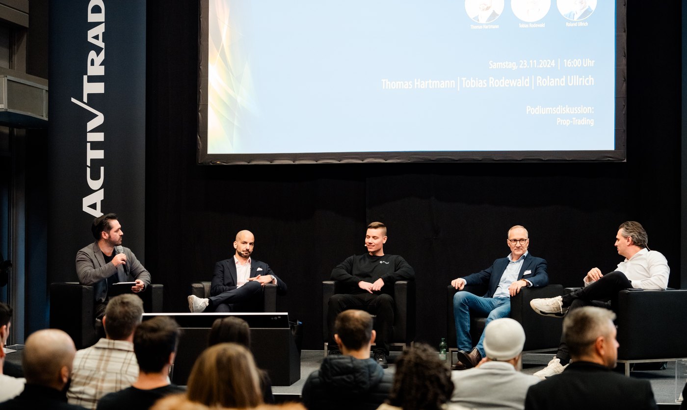 Bühne mit Podiumsteilnehmern und Publikum, auf der Leinwand die Ankündigung der Podiumsdiskussion